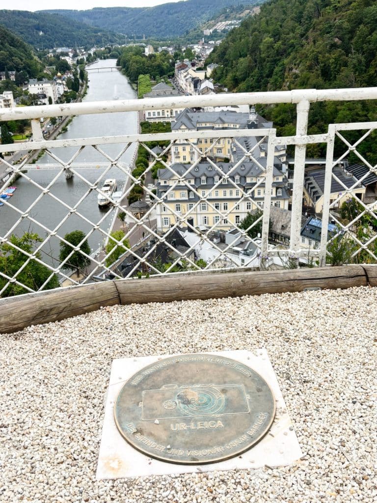 Eine Gedenkplakette für die Ur-Leica und ihren Erfinder Oskar Barnack liegt auf einem kiesbedeckten Aussichtspunkt in Bad Ems, mit Blick über ein weißes Geländer auf den Fluss Lahn und die historische Stadtkulisse.