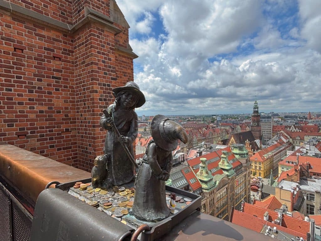 Zwei kleine Bronzefiguren von Zwergen stehen auf einem mit Münzen gefüllten Tablett an der Hexenbrücke in Breslau und blicken über die Dächer der historischen Altstadt unter einem bewölkten Himmel.