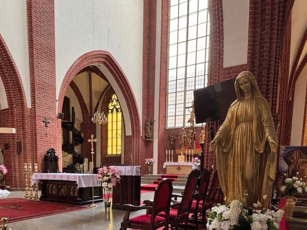 Der Innenraum der Maria-Magdalena-Kirche in Breslau zeigt den Altarbereich mit einer großen goldenen Marienstatue im Vordergrund, roten Samtstühlen und einem Blick durch einen Backsteinbogen auf ein gelbes Buntglasfenster. Der Altar ist mit weißen Blumen und Kerzen geschmückt, während im Hintergrund weitere religiöse Figuren und ein Kruzifix zu sehen sind.
