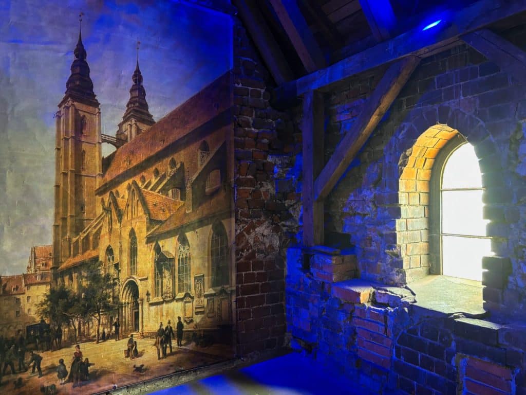 Ein historisches Gemälde der Breslauer Hexenbrücke und der Maria-Magdalena-Kirche hängt an einer rustikalen Backsteinwand in einem Raum mit blauem Licht und einem kleinen Bogenfenster.