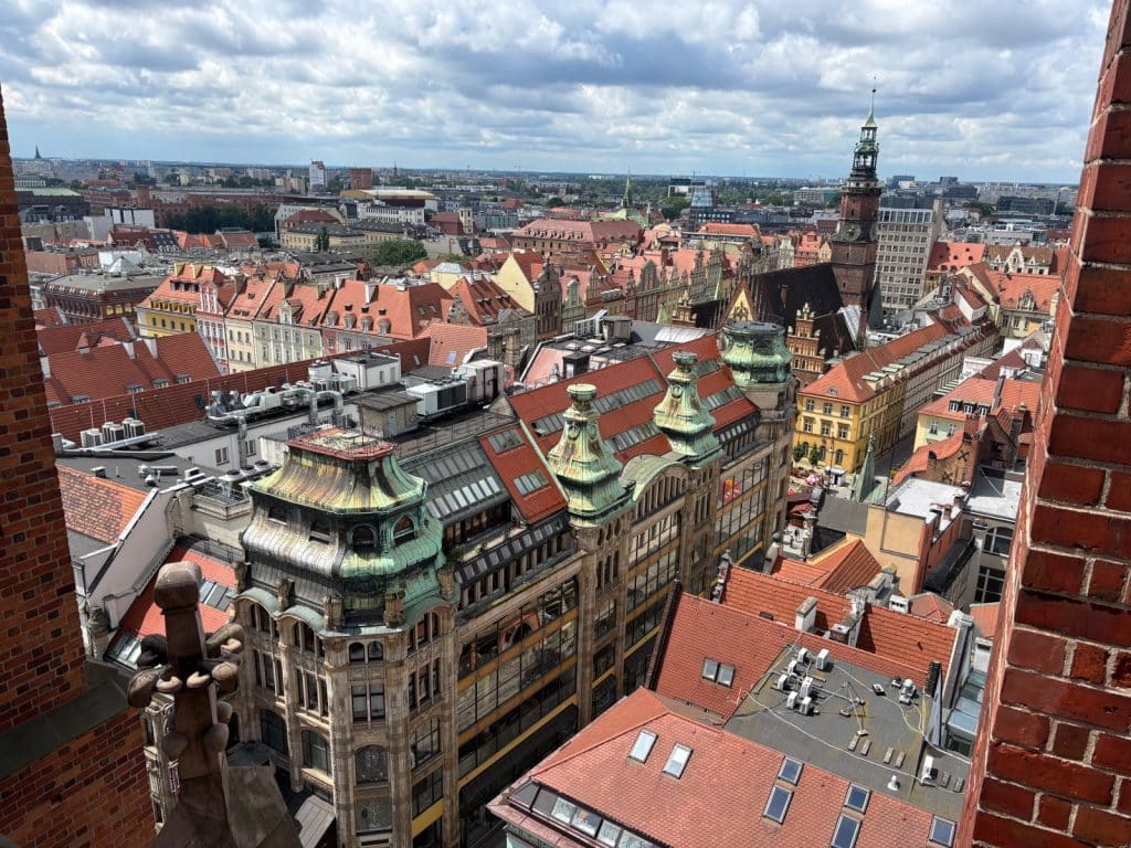 Blick von der Breslauer Hexenbrücke über die historische Altstadt mit ihren charakteristischen roten Ziegeldächern, prachtvollen Bürgerhäusern und dem markanten Turm des Rathauses unter einem bewölkten Himmel.