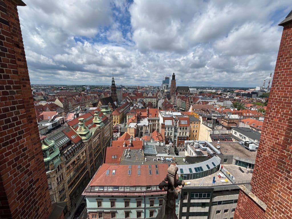 Blick von der Breslauer Hexenbrücke über die historische Altstadt mit ihren roten Ziegeldächern, Kirchtürmen und einer Mischung aus traditioneller und moderner Architektur unter einem bewölkten Himmel.