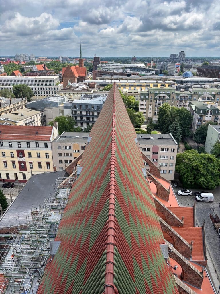Blick von oben auf das steile, mit einem rot-grünen Zickzackmuster gedeckte Dach einer Kirche in Breslau, das direkt auf die Hexenbrücke und das umliegende Stadtpanorama zuführt.