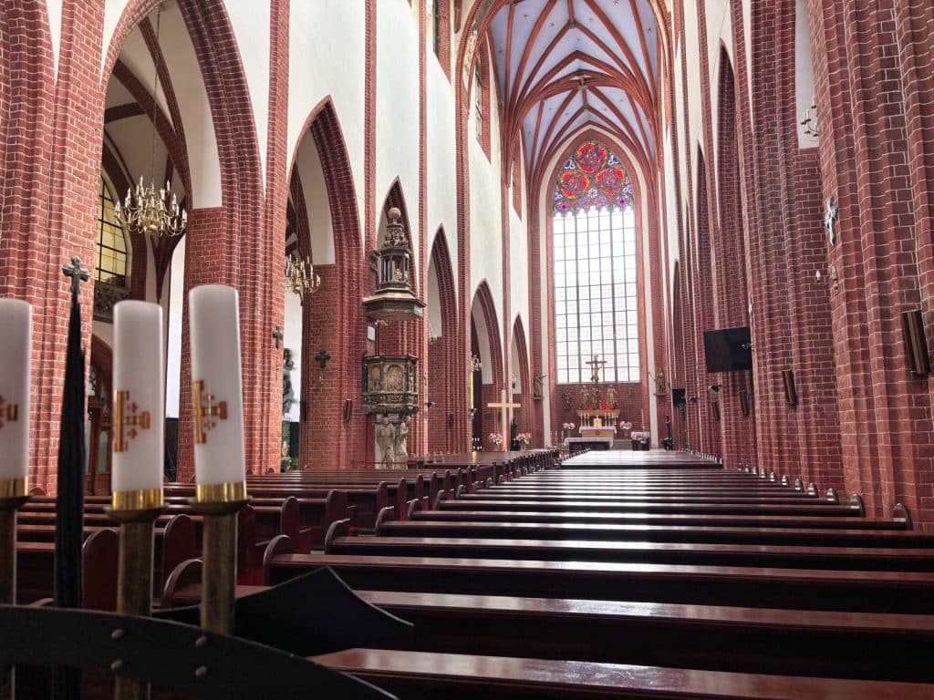 Innenansicht der Maria-Magdalena-Kirche in Breslau mit Blick durch das hohe, backsteinerne Kirchenschiff auf den Altar und ein großes Buntglasfenster, im Vordergrund stehen hölzerne Kirchenbänke und Kerzen.