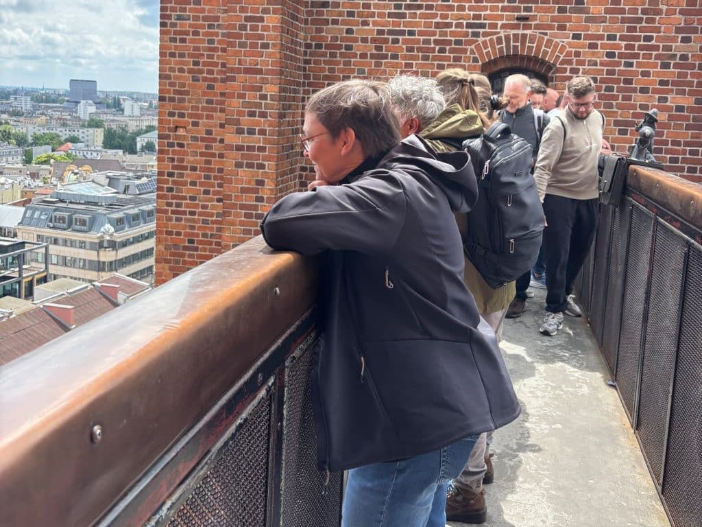 Susanne blickt von der Breslauer Hexenbrücke aus über das Geländer hinunter auf die Stadtlandschaft, während weitere Personen auf dem schmalen Gang der Backsteinkonstruktion stehen.