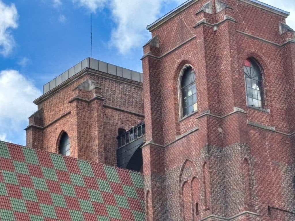 Nahaufnahme der beiden Backsteintürme der Maria-Magdalena-Kirche in Breslau, die durch die hochgelegene, schmale Hexenbrücke miteinander verbunden sind. Im Vordergrund ist ein Teil des Kirchendachs mit einem markanten rot-grün karierten Ziegelmuster zu sehen.