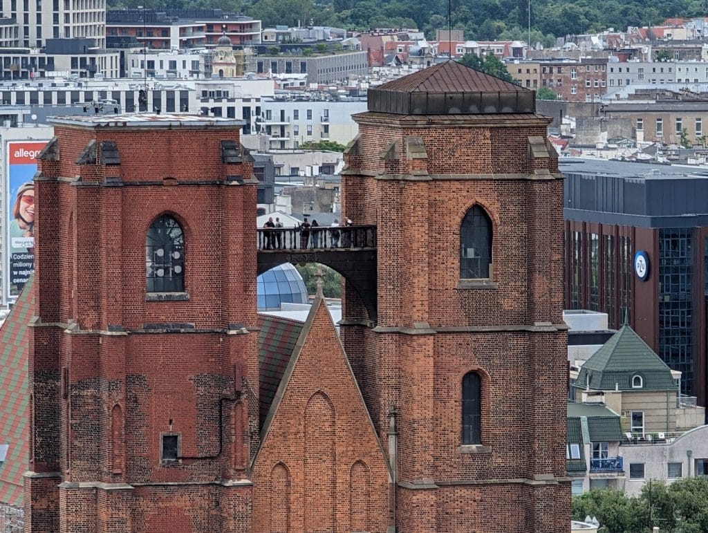 Nahaufnahme der beiden Backsteintürme der Maria-Magdalena-Kirche in Breslau, die durch die hochgelegene Hexenbrücke mit mehreren Personen darauf verbunden sind, vor einer modernen Stadtkulisse.