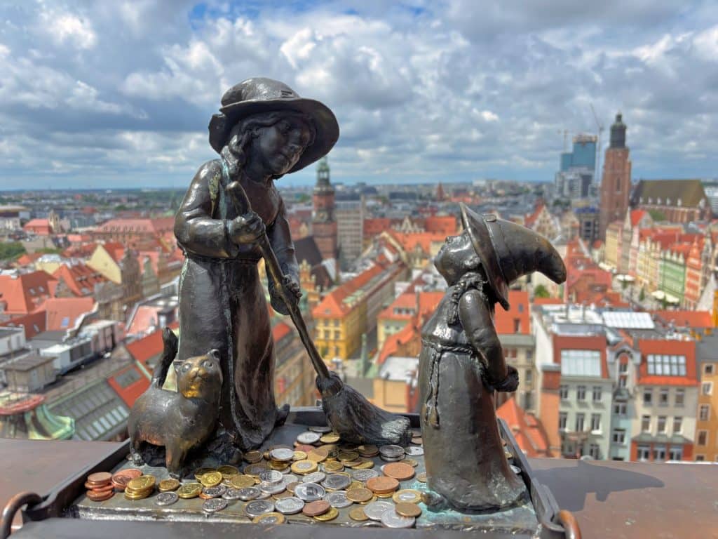 Eine Nahaufnahme von zwei bronzenen Zwergenfiguren, einer Hexe mit Besen und einer Katze, auf der Hexenbrücke in Breslau vor einer weiten Stadtkulisse mit roten Dächern. Zu den Füßen der kleinen Statuen liegen zahlreiche Münzen auf dem Geländer.