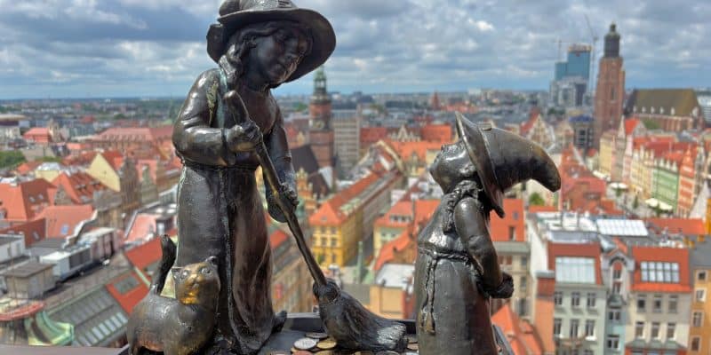 Eine Nahaufnahme von zwei bronzenen Zwergenfiguren, einer Hexe mit Besen und einer Katze, auf der Hexenbrücke in Breslau vor einer weiten Stadtkulisse mit roten Dächern. Zu den Füßen der kleinen Statuen liegen zahlreiche Münzen auf dem Geländer.