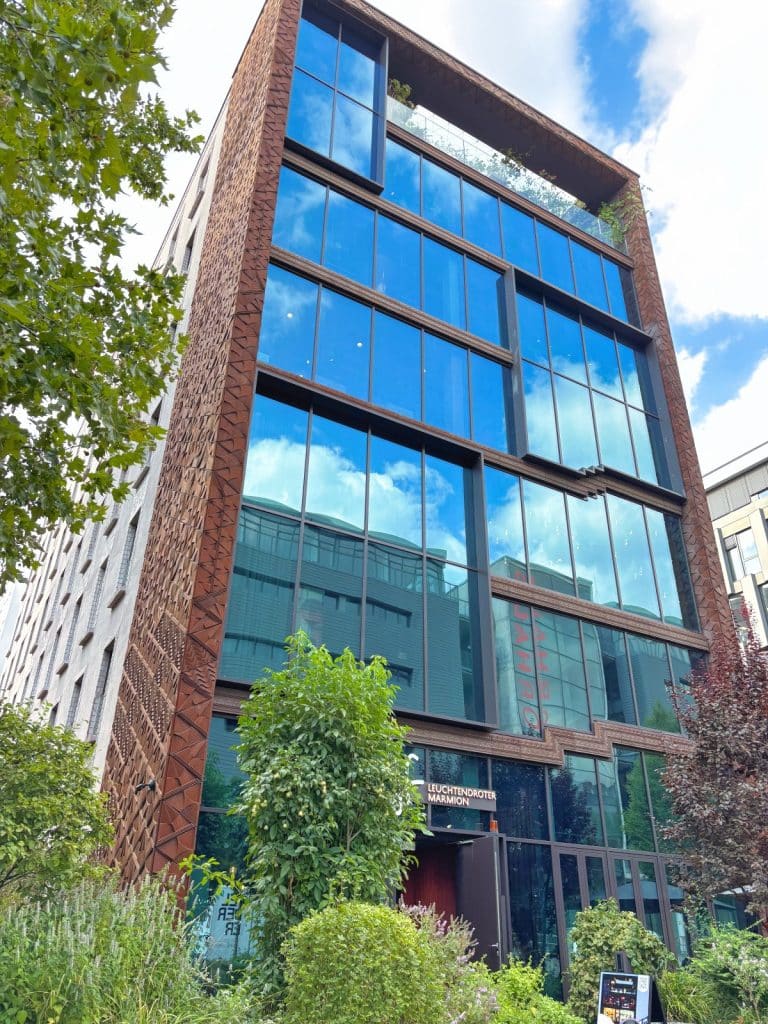 Außenansicht des modernen Hotels lyf EAST FRANKFURT mit einer markanten Fassade aus großen Glasfronten und einer strukturierten, rostbraunen Umrahmung. Im Erdgeschoss befindet sich der Eingang zum Restaurant „Leuchtendroter Marmion“, umgeben von grünen Bäumen und Sträuchern.