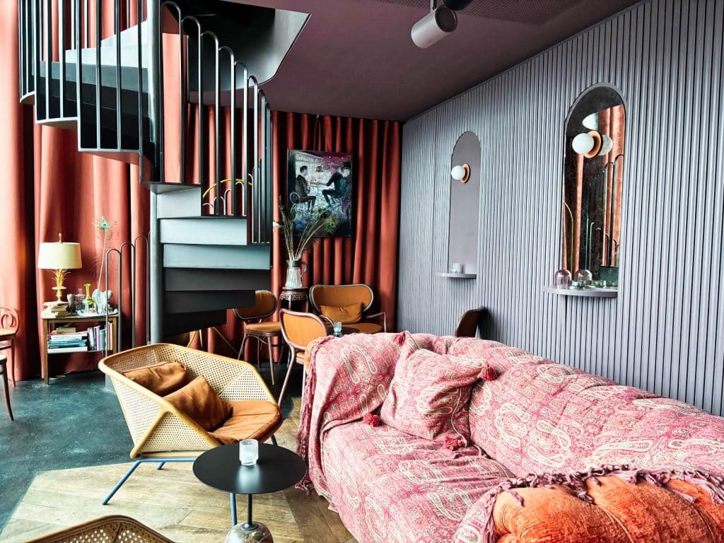 Stilvoll eingerichteter Loungebereich der lyv bar in Frankfurt am Main mit einer markanten schwarzen Wendeltreppe, gemütlichen Sesseln und einem Sofa mit rötlichem Paisley-Muster vor einer grau geriffelten Wand.