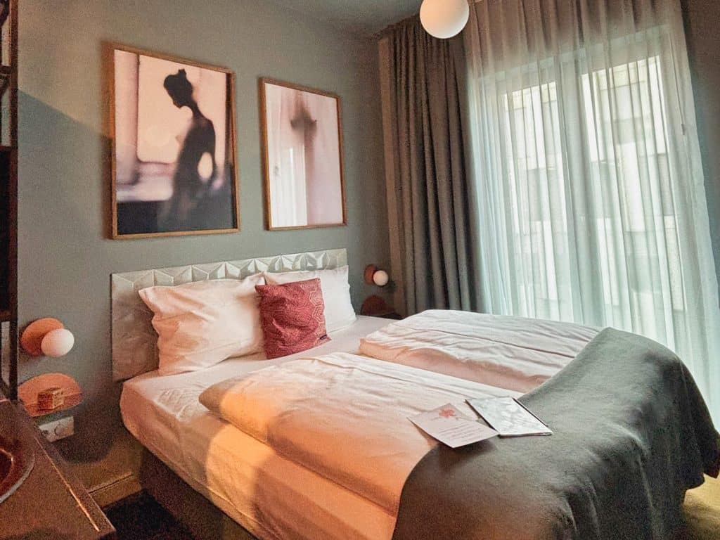 Ein modern eingerichtetes Hotelzimmer im lyf EAST FRANKFURT mit einem Doppelbett, zwei abstrakten Wandbildern über dem gepolsterten Kopfteil und bodentiefen Fenstern mit hellen Vorhängen.