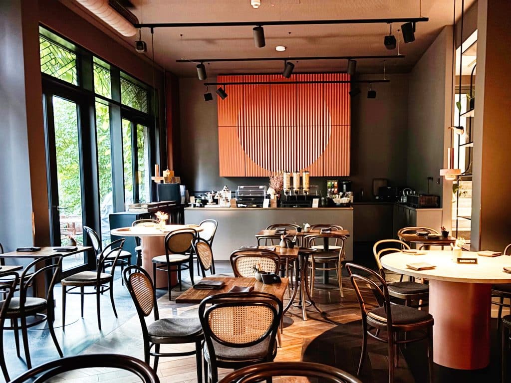 Ein modern gestalteter Frühstücksraum mit runden und eckigen Holztischen, Wiener Geflecht-Stühlen und einem markanten, orangefarbenen Wandkunstwerk hinter der Theke. Durch große Fensterfronten fällt Tageslicht auf den dunklen Parkettboden und die stilvolle Inneneinrichtung.