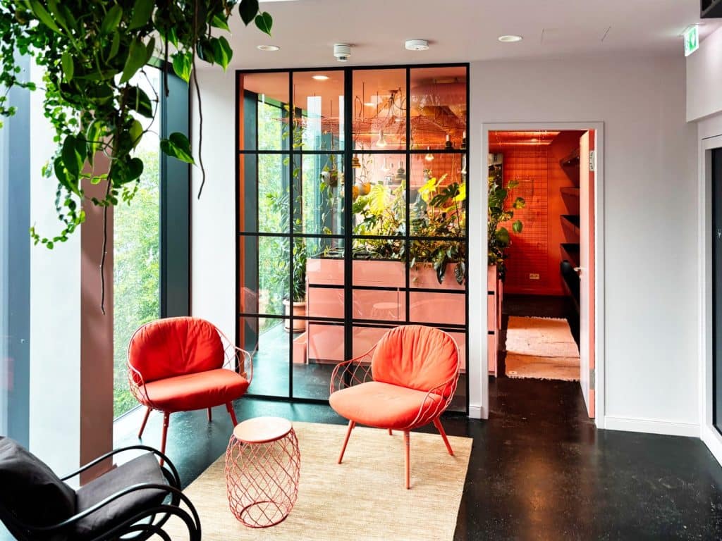 Ein moderner Gemeinschaftsraum Hotel mit zwei korallenfarbenen Sesseln, einem passenden Beistelltisch und einer großen Glasfront, die den Blick auf einen angrenzenden, mit Pflanzen gefüllten Raum freigibt.