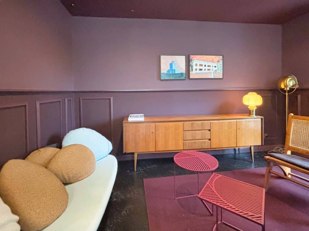 Ein modern gestalteter Gemeinschaftsraum im Hotel in Frankfurt am Main mit dunkelvioletten Wänden, einem hellen Sofa mit runden Kissen, einem Sideboard aus Holz und zwei filigranen, roten Couchtischen auf einem passenden Teppich.