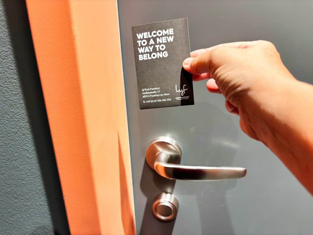 Eine Hand hält eine schwarze Keycard des lyf East Frankfurt Hotels vor eine graue Zimmertür mit silbernem Türgriff. Auf der Karte steht in weißer Schrift „WELCOME TO A NEW WAY TO BELONG“.