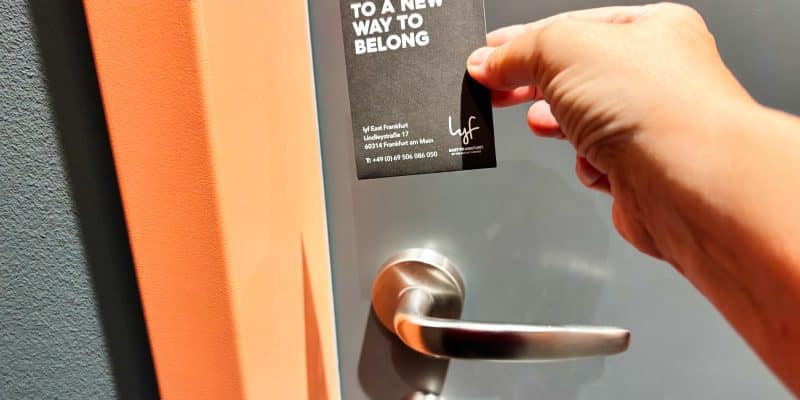 Eine Hand hält eine schwarze Keycard des lyf East Frankfurt Hotels vor eine graue Zimmertür mit silbernem Türgriff. Auf der Karte steht in weißer Schrift „WELCOME TO A NEW WAY TO BELONG“.