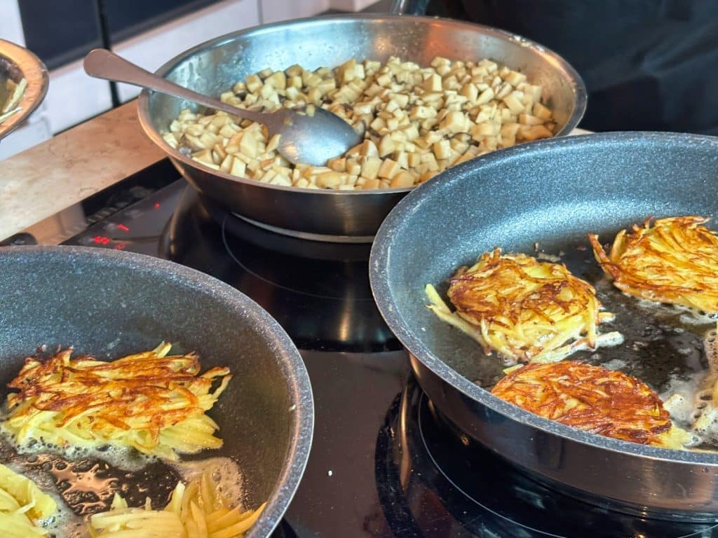 In einer Küche in Frankfurt am Main werden goldbraune Kartoffelpuffer in zwei Pfannen gebraten, während im Hintergrund eine Schüssel mit gewürfelten Pilzen steht.