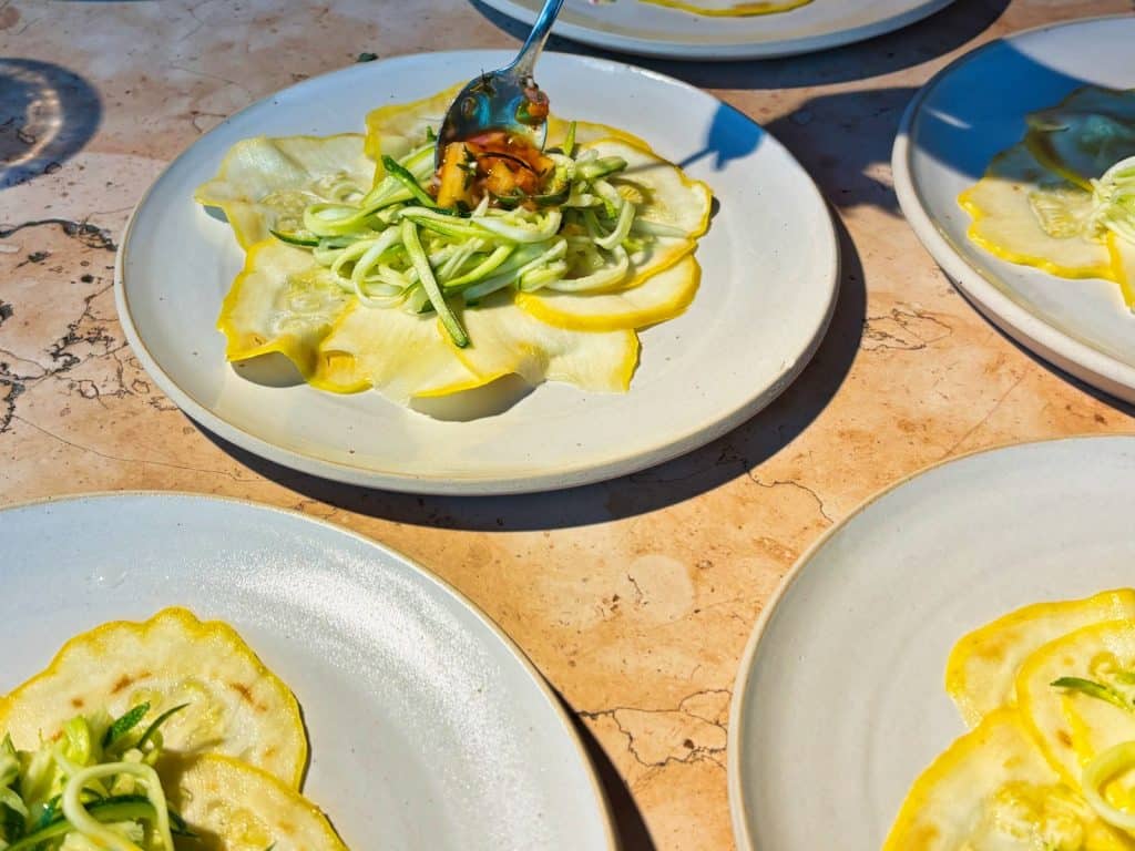 Mehrere weiße Teller mit einer Vorspeise aus dünnen gelben Zucchini-Scheiben und Zucchini-Spaghetti, die bei einem Kochevent von lyf EAST FRANKFURT mit einer Sauce angerichtet werden.