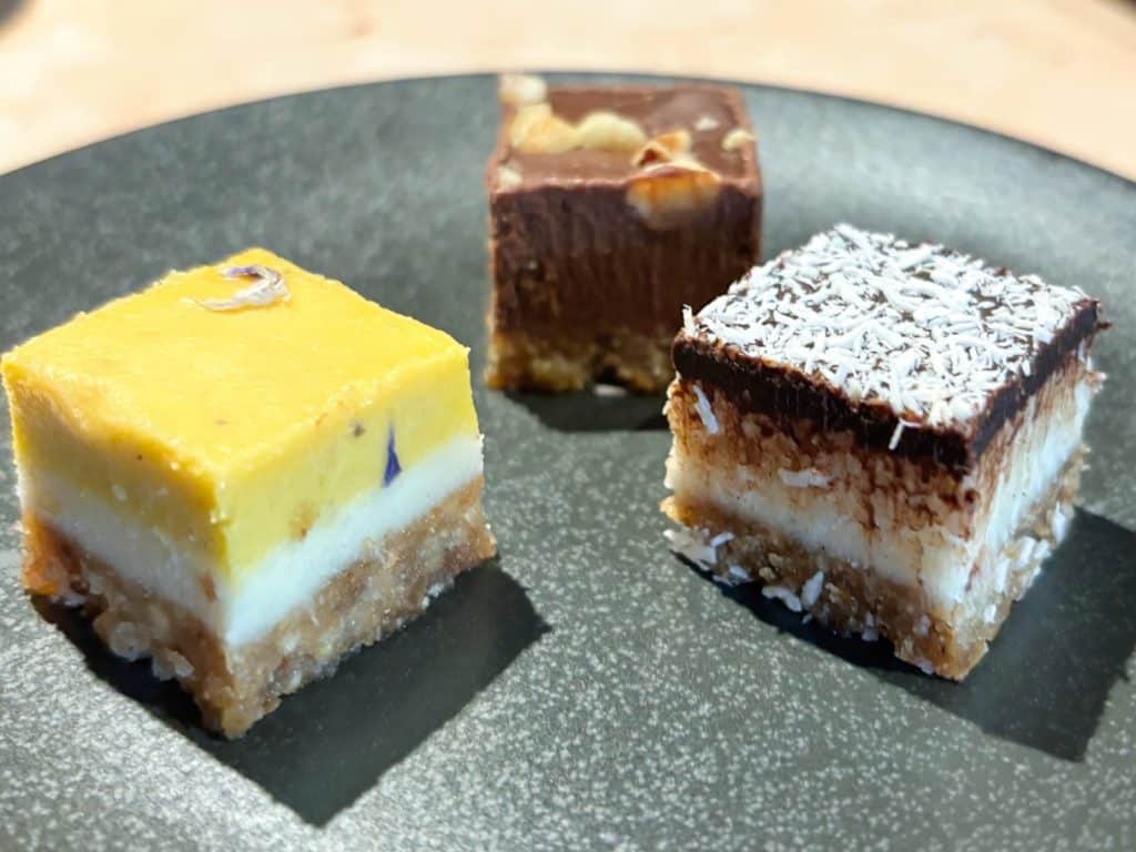 Drei kleine, quadratische Kuchenstücke von lyf EAST FRANKFURT, darunter eine gelbe Zitronen-Schnittte, ein Schokowürfel mit Nüssen und ein Kokos-Schoko-Riegel, sind auf einem dunklen Teller angerichtet.