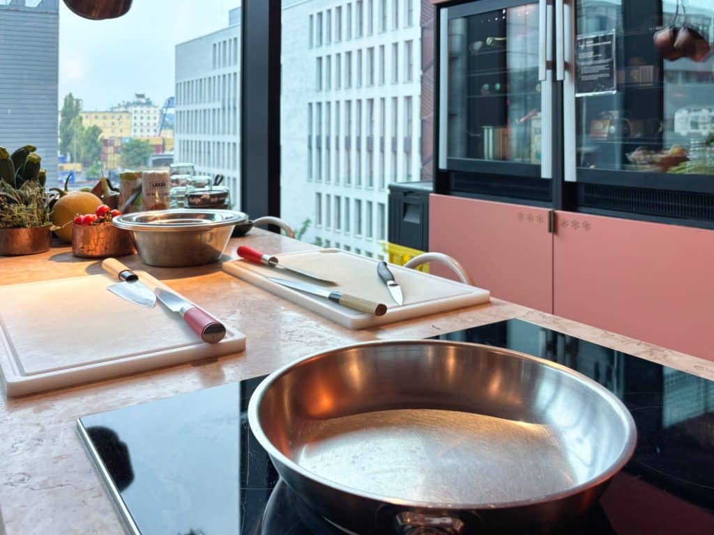 In einer modernen Küche im lyf EAST FRANKFURT liegen Messer und Schneidebretter neben einer Edelstahlpfanne auf dem Herd bereit zum Kochen, während der Blick durch das Fenster auf die Stadt fällt.
