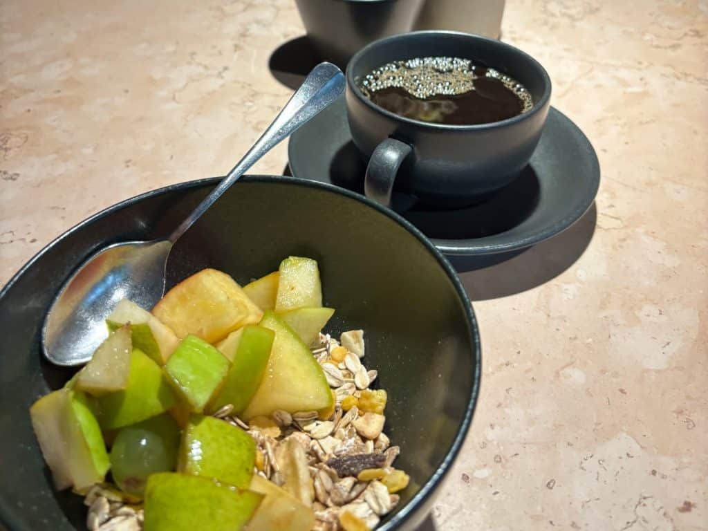 Ein Frühstück mit einer Schale Müsli, das mit frischen Apfelstücken und Weintrauben garniert ist, steht neben einer Tasse schwarzem Kaffee auf einem hellen Steintisch in Frankfurt am Main.