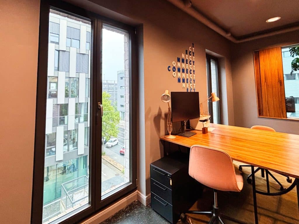 Ein moderner Arbeitsplatz im lyf EAST FRANKFURT in Frankfurt am Main mit einem großen Holzschreibtisch, einem Monitor und einer Wandgestaltung aus dem Wort „CONNECT“ neben einem bodentiefen Fenster mit Blick auf Stadtgebäude.