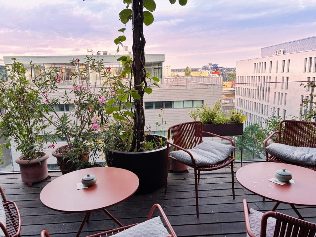 Die Dachterrasse der Bar in lyf EAST FRANKFURT am Main bei Abenddämmerung mit runden Tischen, modernen Sesseln und üppigen Topfpflanzen vor der städtischen Kulisse.