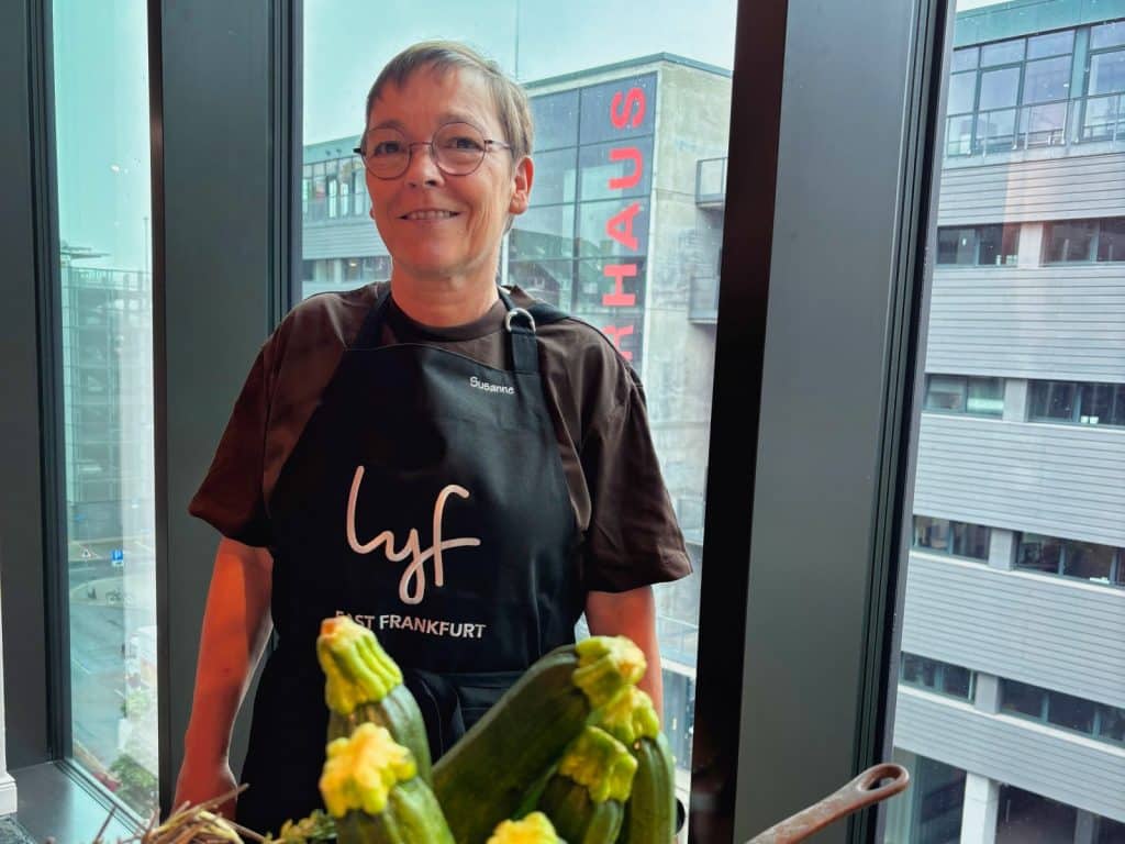 Susanne lächelt in einer schwarzen Schürze mit der Aufschrift „lyf EAST FRANKFURT“, während sie hinter einem Korb mit frischen Zucchini vor einem Fenster mit Blick auf ein städtisches Gebäude steht.