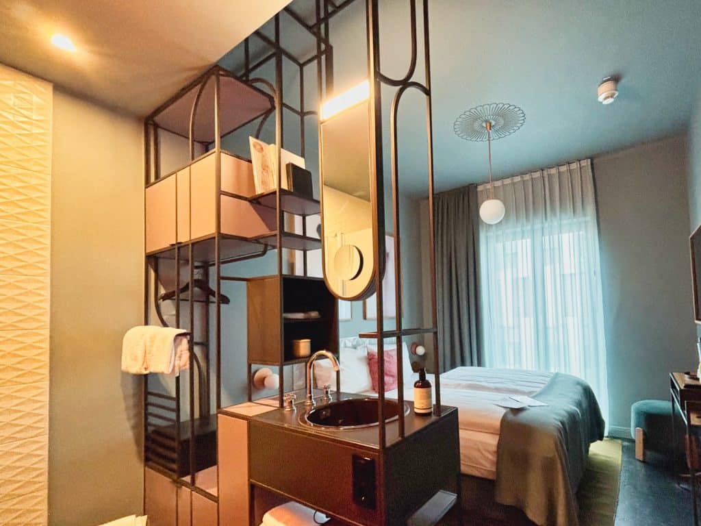 Ein modern gestaltetes Zimmer im lyf EAST FRANKFURT mit einem offenen Badezimmerbereich, der durch ein schwarzes Metallregalsystem mit integriertem Waschbecken und Spiegel vom Schlafbereich getrennt ist.