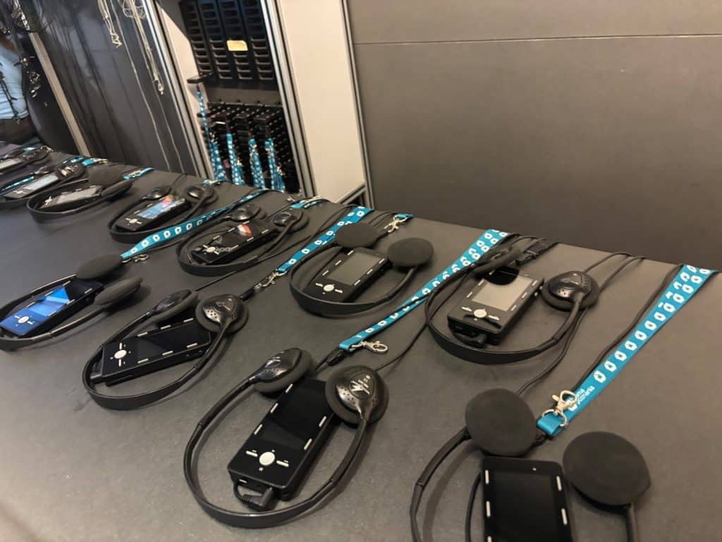 Mehrere schwarze Audioguides mit Kopfhörern und blau gemusterten Tragebändern liegen ordentlich aufgereiht auf einem grauen Tisch vor einem Lagerschrank.