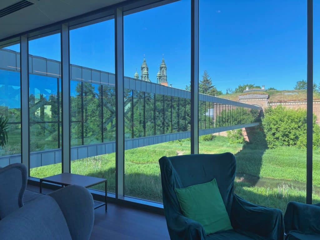Blick aus einem modernen Innenraum mit Sesseln durch eine große Fensterfront auf die gläserne Brama-Brücke in Posen, die über eine Grünfläche zu einem historischen Backsteingebäude führt, mit den Türmen der Posener Kathedrale im Hintergrund.
