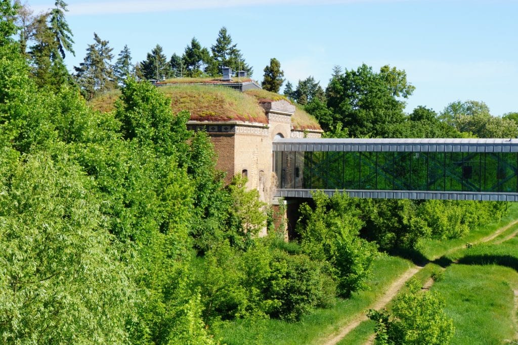 Ein historisches Backsteingebäude mit grasbewachsenem Dach an der Brama Poznańska (Posener Tor) ist durch einen modernen, verglasten Übergang über das Flussufer hinweg mit der anderen Seite verbunden.