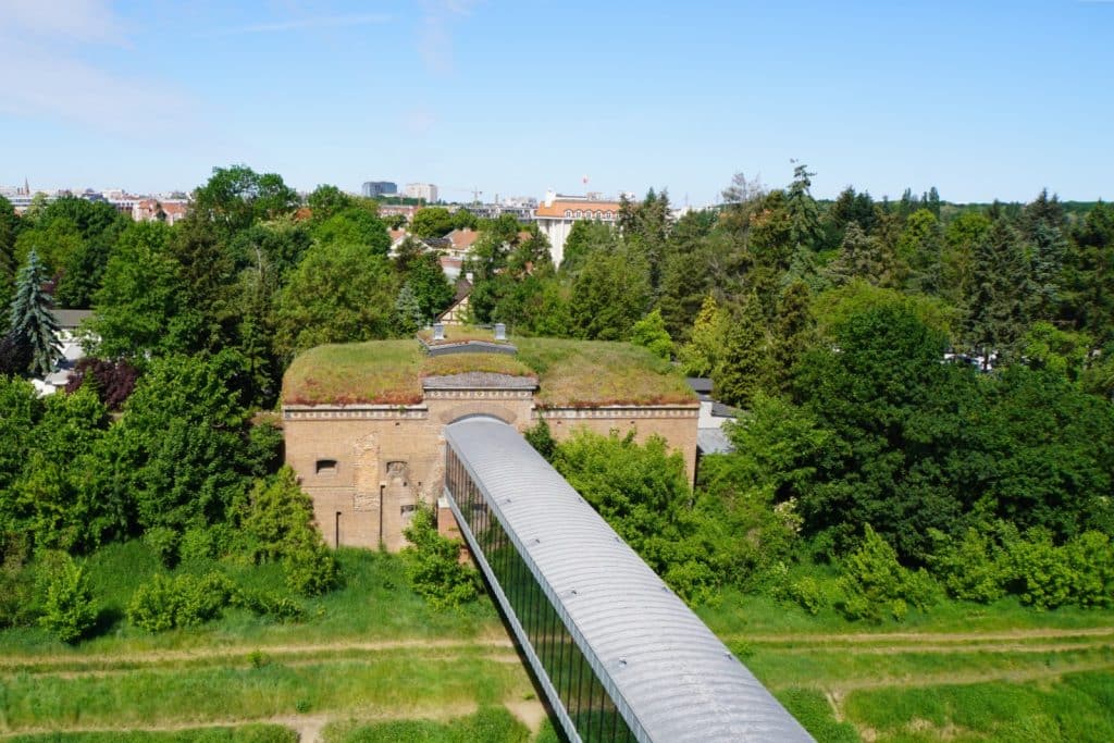 Blick vom Dach auf die historische Brama Poznańska (Posener Tor) in Posen, die durch eine moderne, verglaste Brücke über eine Grünanlage mit dem Hauptgebäude verbunden ist.