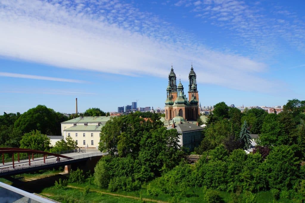 Blick vom Dach des interaktiven Zentrums Brama Poznania auf den Posener Dom, der hinter dichten grünen Bäumen und einer roten Bogenbrücke unter einem weiten blauen Himmel aufragt.