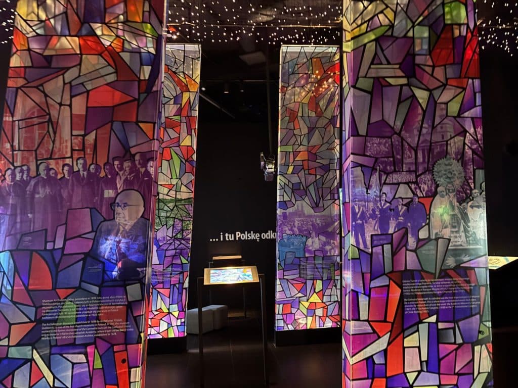 Ein Ausstellungsraum im interaktiven Zentrum Porta Posnania (Brama Poznania) in Posen, in dem beleuchtete Säulen mit bunten Mustern aus Glasbruchstücken und historischen Schwarz-Weiß-Fotografien stehen. Die Glasmosaike leuchten in kräftigen Farben und zeigen Porträts sowie Gruppenszenen vor einem dunklen Hintergrund mit Sternenlichtern an der Decke.