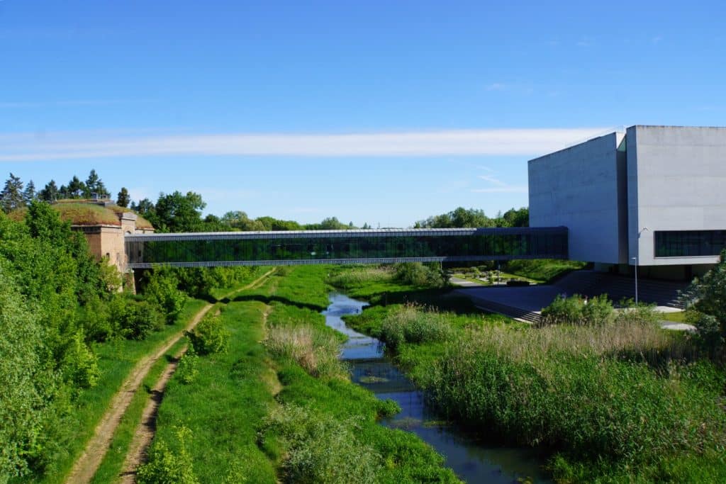 Eine moderne, verglaste Fußgängerbrücke überspannt das grüne Ufer der Cybina und verbindet das historische Brama Poznania mit dem modernen Hauptgebäude des interaktiven Zentrums in Posen. Das Bild zeigt den Kontrast zwischen der alten Backsteinarchitektur und dem minimalistischen Betonbau unter einem klaren blauen Himmel.