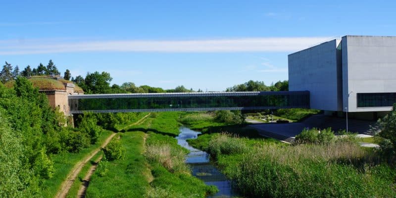Eine moderne, verglaste Fußgängerbrücke überspannt das grüne Ufer der Cybina und verbindet das historische Brama Poznania mit dem modernen Hauptgebäude des interaktiven Zentrums in Posen. Das Bild zeigt den Kontrast zwischen der alten Backsteinarchitektur und dem minimalistischen Betonbau unter einem klaren blauen Himmel.
