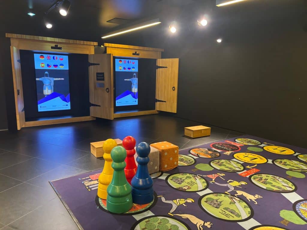 Ein interaktiver Ausstellungsraum im Brama Poznania zeigt eine spielerische Geschichtsvermittlung mit einem großen Boden-Brettspiel, riesigen bunten Spielfiguren und digitalen Bildschirmen in holzgerahmten Nischen.