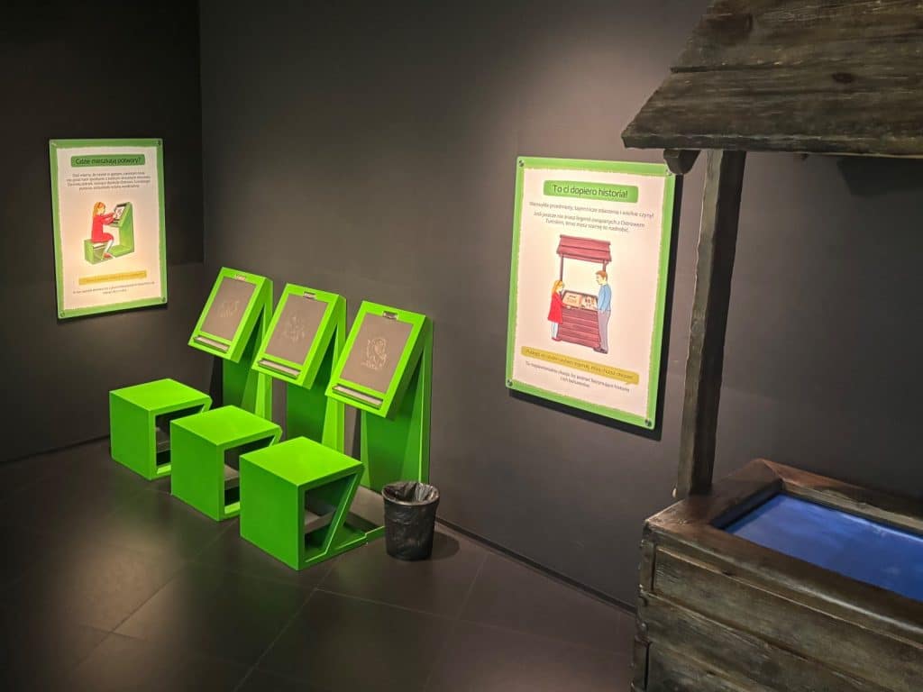 Eine Kinderstation im interaktiven Zentrum Brama Poznania mit drei grünen Sitzplätzen vor Zeichentafeln, erklärenden Postern an der dunklen Wand und einem hölzernen Brunnenmodell im Vordergrund.
