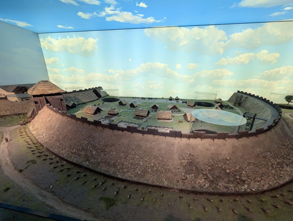 Ein detailliertes Modell der historischen Burg von Posen im Brama Poznania zeigt eine befestigte Siedlung mit Holzhäusern, die von einem hohen Erdwall und Palisaden umgeben ist. Das Modell steht vor einer Hintergrundwand mit gemaltem blauem Himmel und weißen Wolken, während gläserne Zylinder Einblicke in archäologische Schichten ermöglichen.