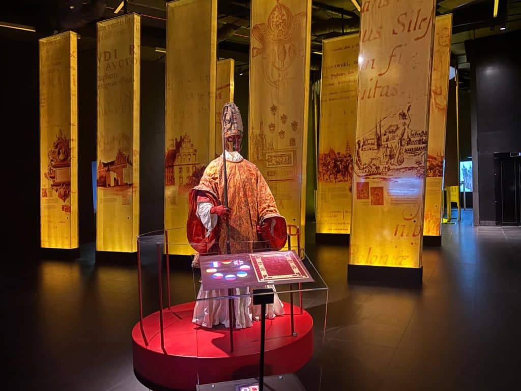 Eine Museumsausstellung im Brama Poznania in Posen zeigt ein historisches Bischofsgewand auf einer roten Plattform, umgeben von hohen, beleuchteten Glasstelen mit lateinischen Texten und Illustrationen. Das Gewand besteht aus einer goldenen Kasel, einer Mitra und einem Stab, während Informationstafeln im Vordergrund Details zur Geschichte vermitteln.