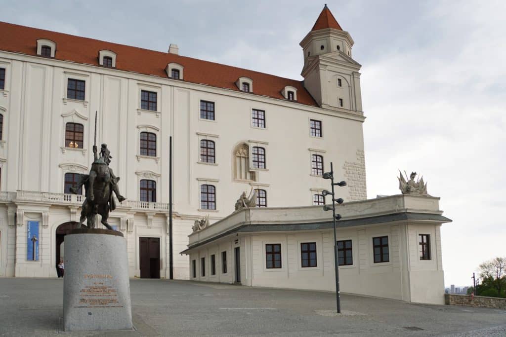 Die weiße Bratislava Burg mit ihren markanten roten Dächern und einem Ecktürmchen ragt hinter einem Reiterdenkmal auf einem gepflasterten Vorplatz empor. Im Vordergrund steht ein flaches, weißes Nebengebäude mit einer Balustrade, auf der Skulpturen thronen.