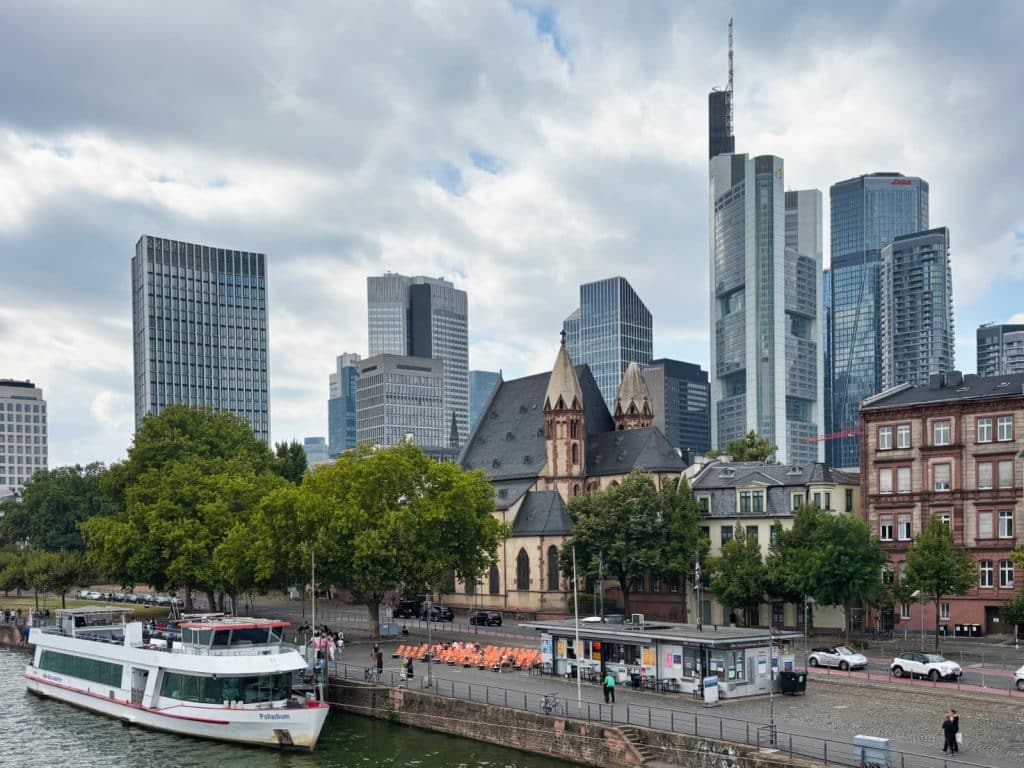 Blick auf die Frankfurter Skyline am Main mit modernen Wolkenkratzern wie dem Commerzbank Tower hinter der historischen Dreikönigskirche, während im Vordergrund ein Ausflugsschiff am Flussufer anlegt.