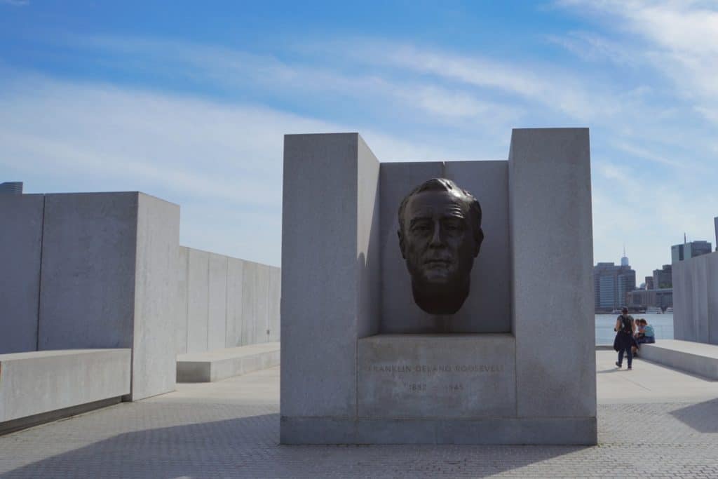Ein monumentaler Granitblock mit der Bronzebüste von Franklin D. Roosevelt im Four Freedoms Park in New York, umgeben von weiteren hellen Steinquadern unter einem blauen Himmel.
