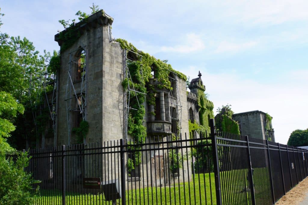 Die Ruinen des ehemaligen Roosevelt-Krankenhauses auf Roosevelt Island in New York zeigen eine verfallene Steinstruktur, die stark mit Efeu bewachsen und von einem schwarzen Metallzaun sowie Gerüsten umgeben ist.