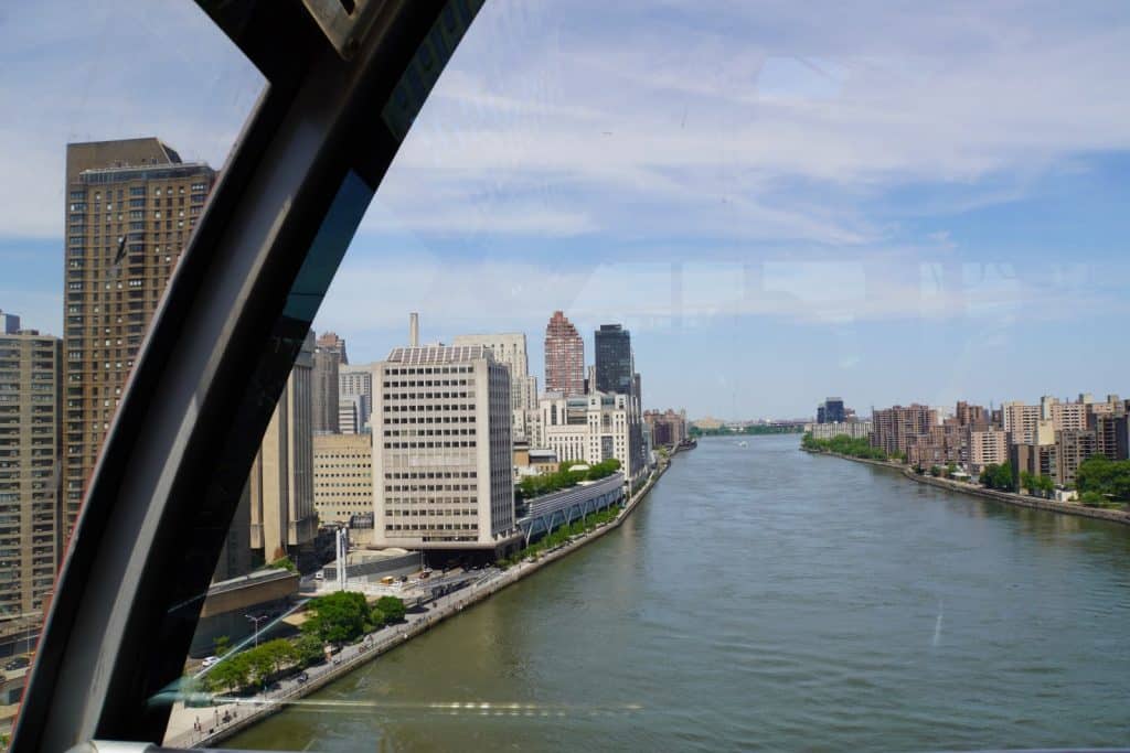 Blick aus dem Fenster der New Yorker Roosevelt Island Seilbahn über den East River auf die Skyline von Manhattan und Roosevelt Island unter einem leicht bewölkten Himmel.