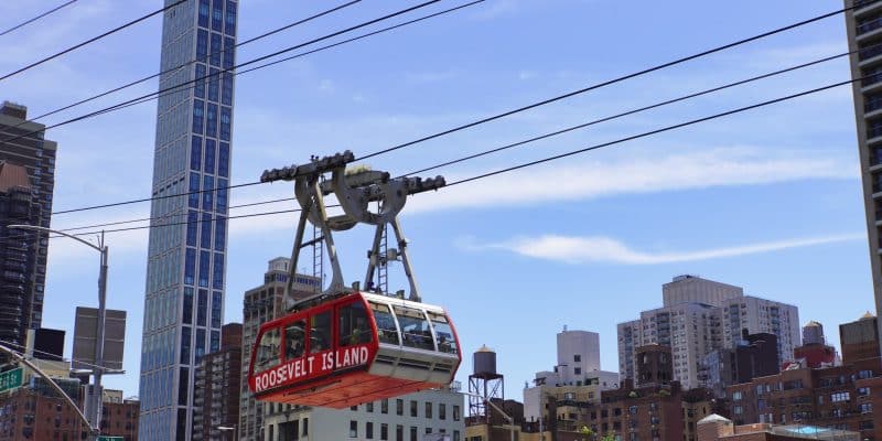 Eine rote Kabine der Roosevelt Island Tramway schwebt an dicken Drahtseilen vor der Skyline von New York City mit zahlreichen Hochhäusern unter einem blauen Himmel. Die Seilbahn ist nah im Profil zu sehen und trägt die weiße Aufschrift „ROOSEVELT ISLAND“.