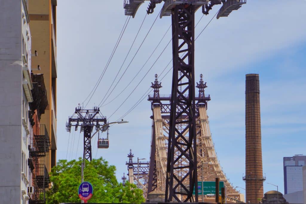 Blick auf die Roosevelt Island Tramway in New York City mit einer roten Seilbahnkabine, die vor der markanten Stahlkonstruktion der Queensboro Bridge schwebt.