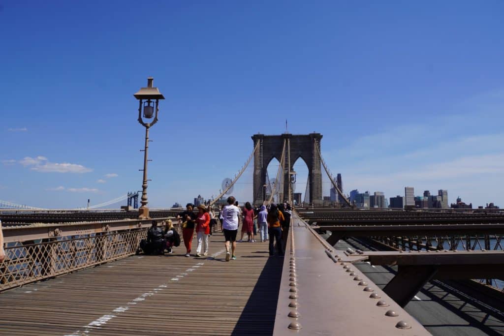 Zahlreiche Menschen spazieren bei strahlendem Sonnenschein über den hölzernen Fußgängerweg der Brooklyn Bridge in Richtung eines der markanten neugotischen Steinpfeiler. Links im Bild ragt eine historische Straßenlaterne empor, während im Hintergrund die Skyline von Manhattan unter einem klaren blauen Himmel zu sehen ist.